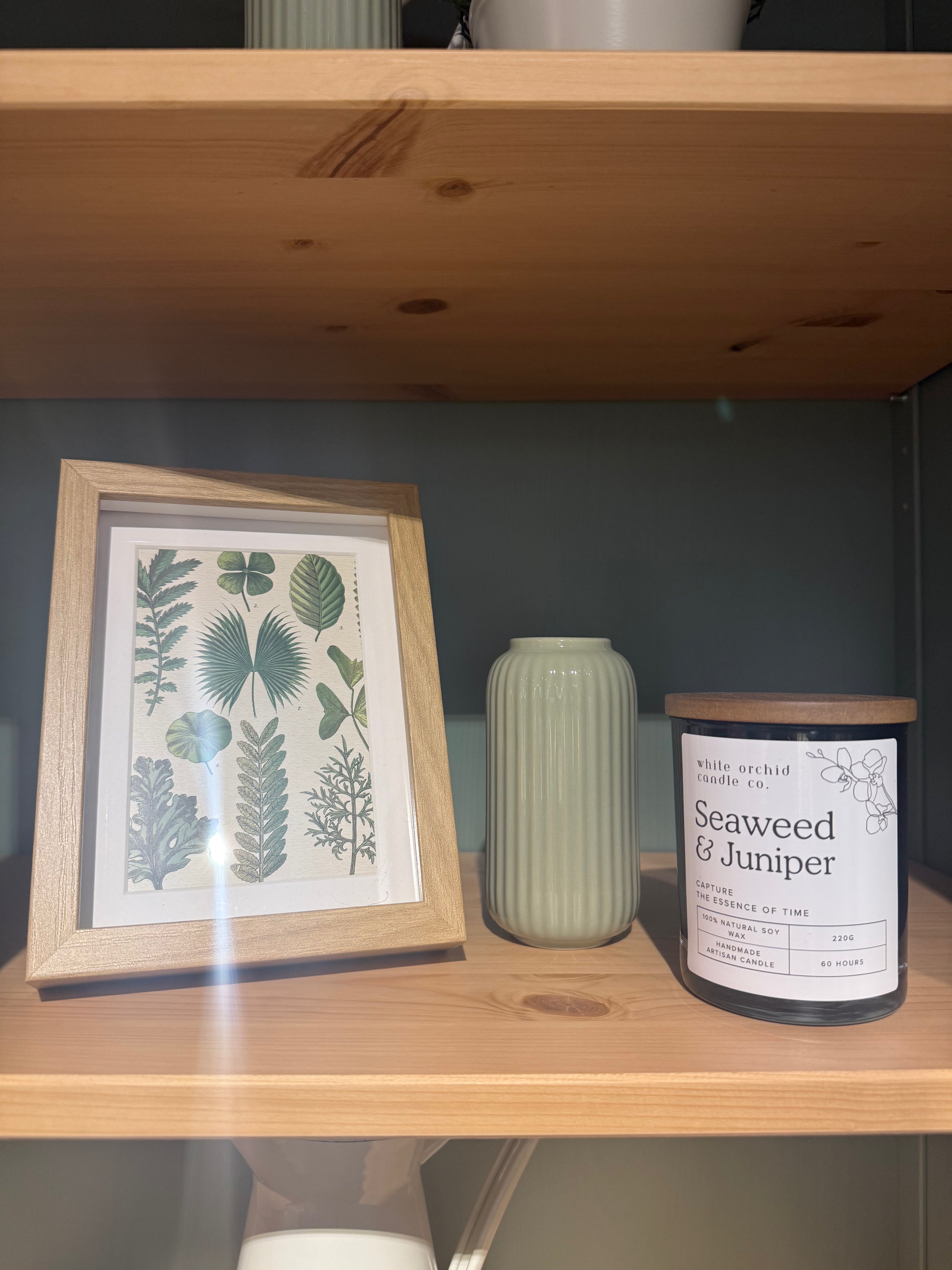 Botanical wall art, a green vase, and a Seaweed & Juniper scented candle styled on a wooden shelf.